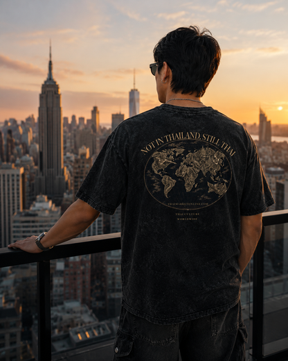 A man stands on a rooftop balcony at sunset, viewed from behind, wearing a faded black graphic t-shirt with a gold world map design and the text 'MOTAN THAILAND STILL THA'. The background shows a city skyline with the Empire State Building under a warm orange sky.