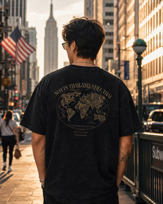 A man viewed from behind wearing a black graphic t-shirt with a gold world map design and the text 'NOT IN THAILAND STILL HERE' on the back, standing on a city street.