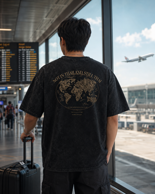 A man stands with his back to the camera in an airport terminal, wearing a faded black graphic t-shirt with a gold world map design and the text 'NOT IN THAILAND, STILL THAI'. He is holding a black suitcase next to large windows showing an airplane flying overhead.